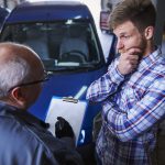 Älterer Mann mit Aktenordner spricht in einer Werkstatt mit einem besorgten Autobesitzer, während im Hintergrund dessen Auto bei der TÜV Prüfung inspiziert wird