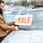 Person befestigt einen Zu verkaufen Zettel an der Windschutzscheibe eines Gebrauchtwagens auf einem Parkplatz